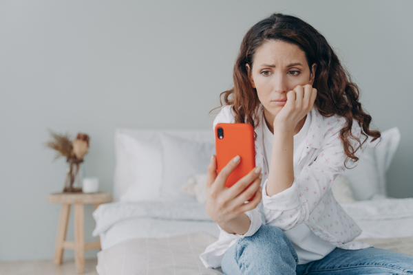 A woman sits on the couch staring at an orange phone with a puzzled look on her face.