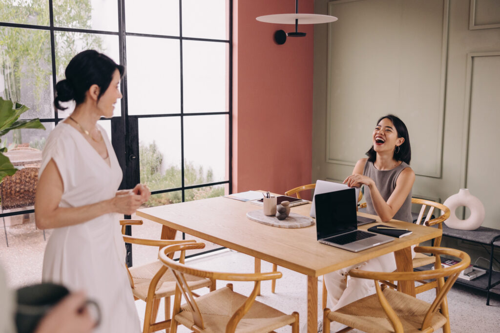 Two women are laughing as one stands near a table and the other sits.