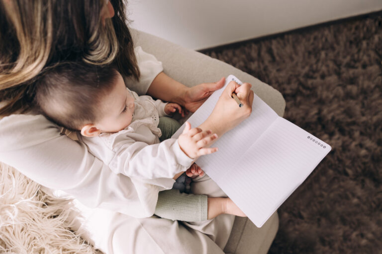 Mother multitasking with a baby on her lap while writing in a notebook, symbolizing the flexibility of growing a doula business in small, manageable time blocks.