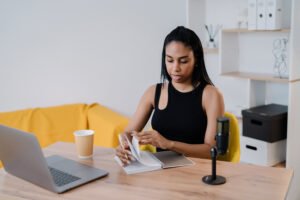 Black woman sits at a desk and flips through a notebook with a coffee cup, computer, and podcast microphone at the table.