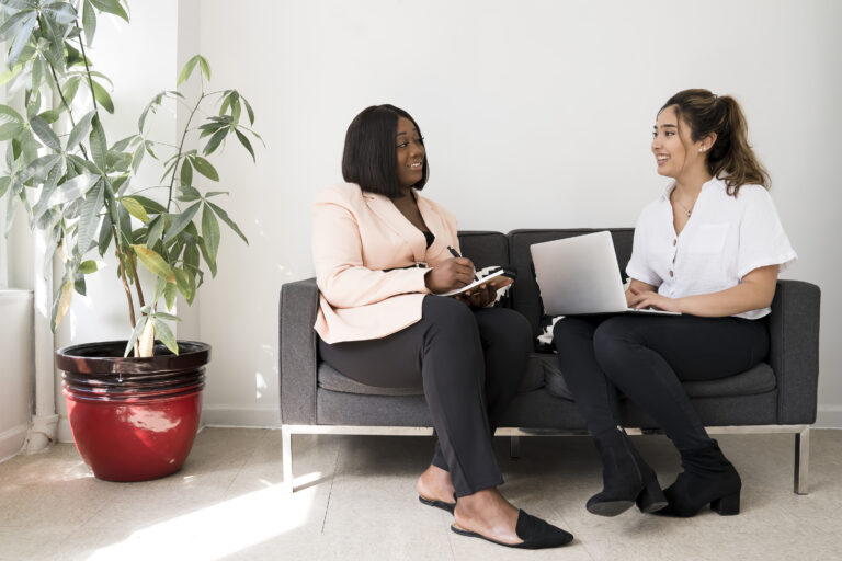 Two women sit on a couch and discuss work.