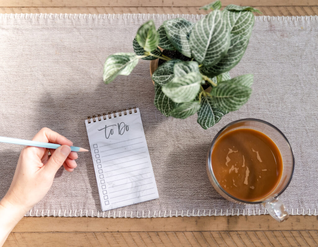 A hand hovers over a list that says to do, near a cup of coffee and a plant.