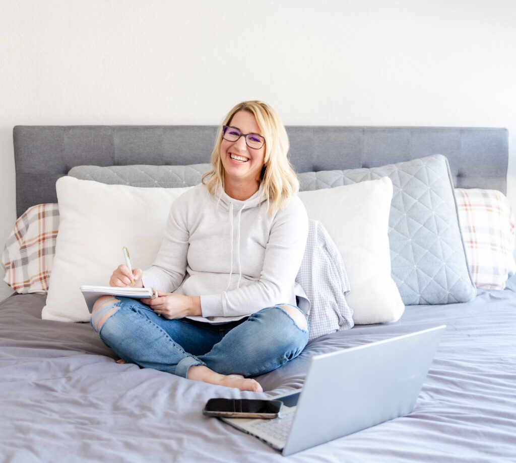 A doula in a white shirt and blue jeans sits cross legged on her bed writing on a pad of paper.