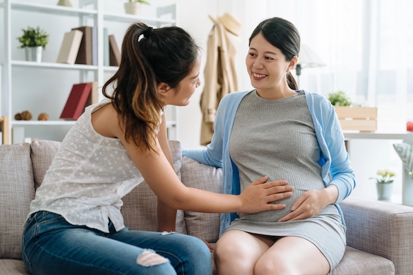 smiling pregnant woman communicates with girl friend indoors. young future motherhood lady showing her belly with baby expect bestie hands touching feeling. two female sitting on sofa chatting home