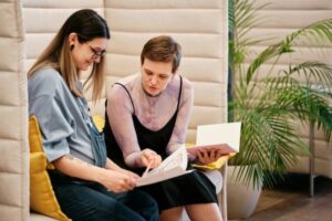 Two women sit in chairs and talk as one points to a book.