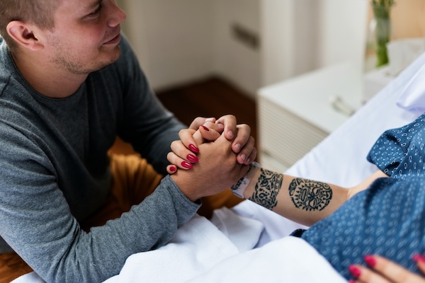 A man holds the hand of a pregnant person in a hospital gown with tattoos.