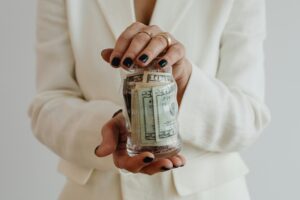 A woman in a white shirt holds up a jar with paper cash in it.