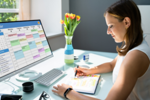 Woman sitting at a desk writing in a planner while looking at a calendar on a computer screen, representing how birth doulas plan their monthly client schedule.