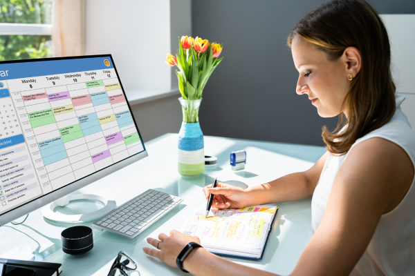 Woman sitting at a desk writing in a planner while looking at a calendar on a computer screen, representing how birth doulas plan their monthly client schedule.