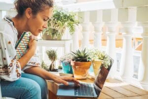 A doula works on her laptop at a sunlit balcony table surrounded by potted plants, paused mid-thought with one hand on the keyboard.