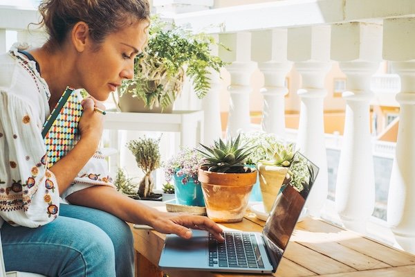 A doula works on her laptop at a sunlit balcony table surrounded by potted plants, paused mid-thought with one hand on the keyboard.
