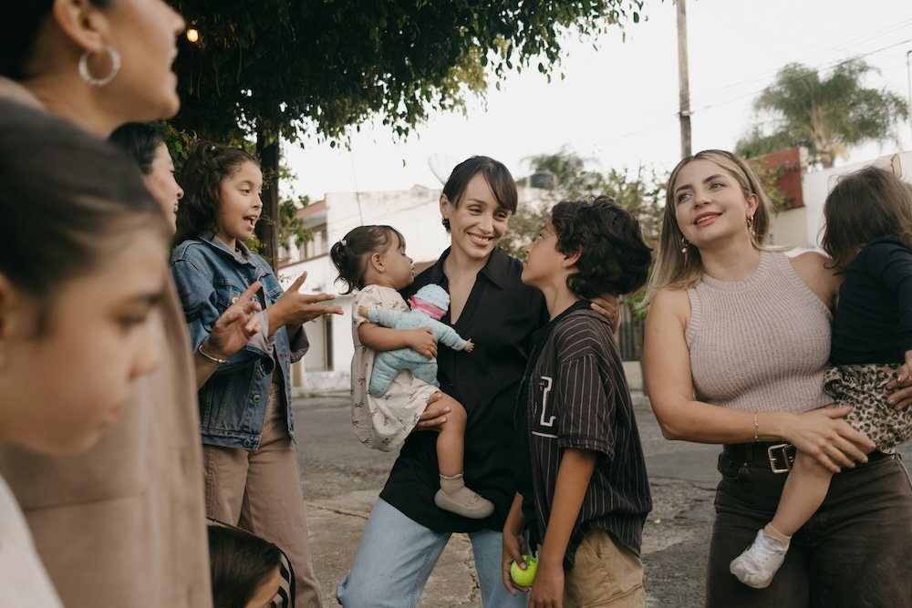 A group of women and children standing close together on a neighborhood street in evening light, two women each carrying a toddler as older kids gather around them, laughing.
