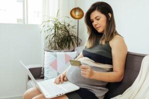 A pregnant woman sits with a computer and a credit card.