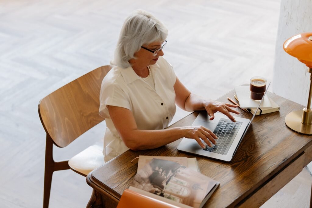 A silver-haired doula types on a laptop at a wooden desk.