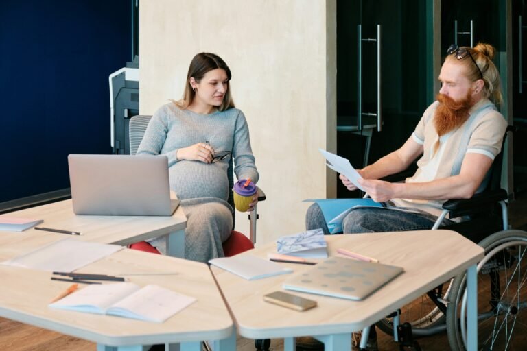 A pregnant person holding a coffee cup talks with a partner in a wheelchair who is reading a printed page at an office table covered with a laptop, notebooks and pencils.