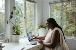 A person with long braids and glasses taps a laptop screen at a bright window desk, with a calculator, an envelope, and plants nearby, checking how their doula business appears in AI search.